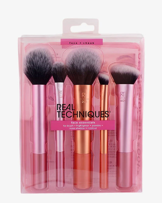 A 5-piece makeup brush set in its pink translucent plastic packaging, featuring pink and rose gold handles and black bristles. The brand 'Real Techniques' is clearly visible on the front of the packaging.